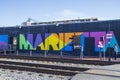 A colorful wall mural with the word âMariettaâ along the railroad tracks with people standing on the corner in Marietta